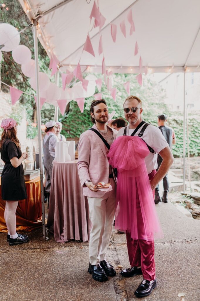 A man in a pink suit jacket and a man with hot pink tuile in his leather bands stand together.