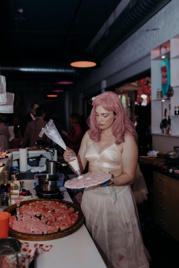 A woman in a white dress with pink hair frosts white frosting onto a plate.