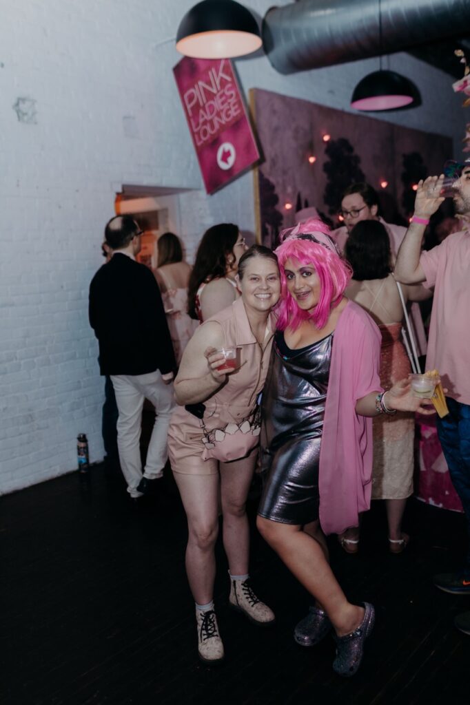 A woman in a silver dress and pink wig stands with a woman in a pink romper.