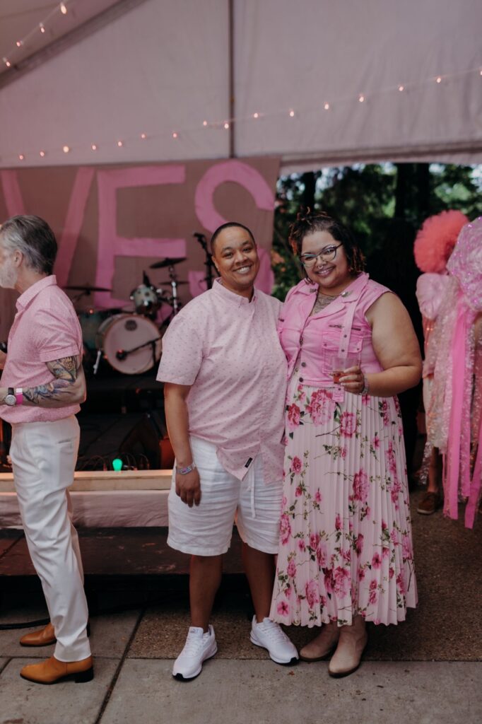 A woman in a pink button up stands with a woman in a pink rose dress.