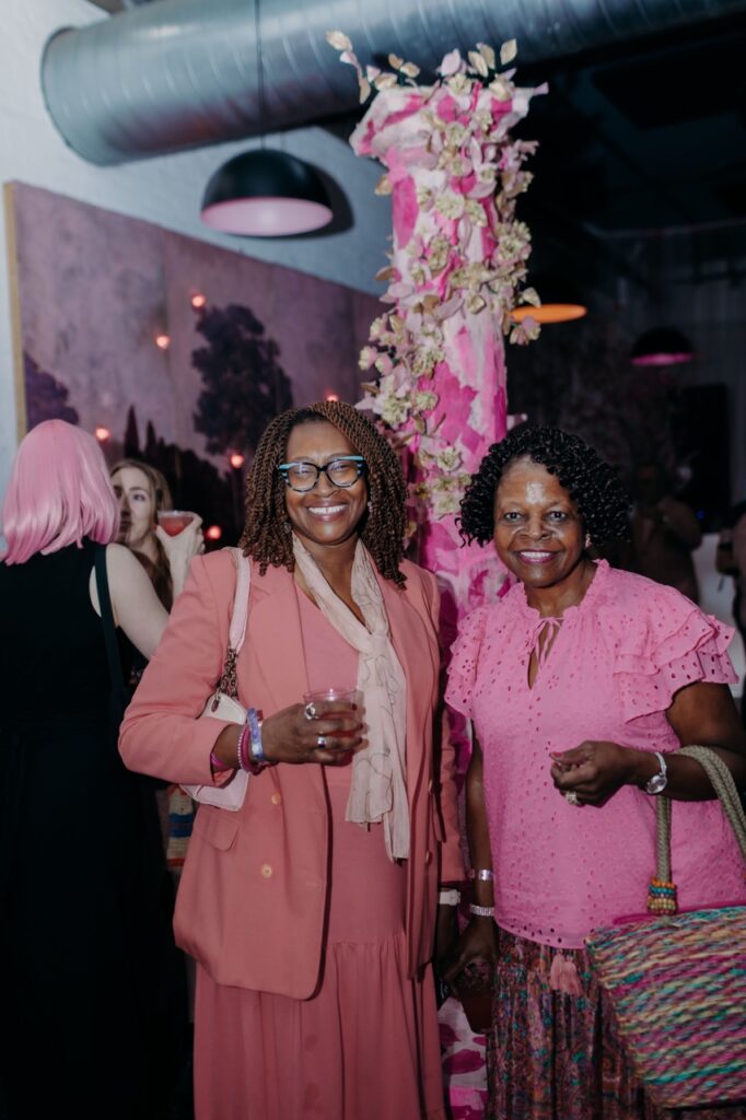 Two women in pink stand in front of a pink, flower covered column.
