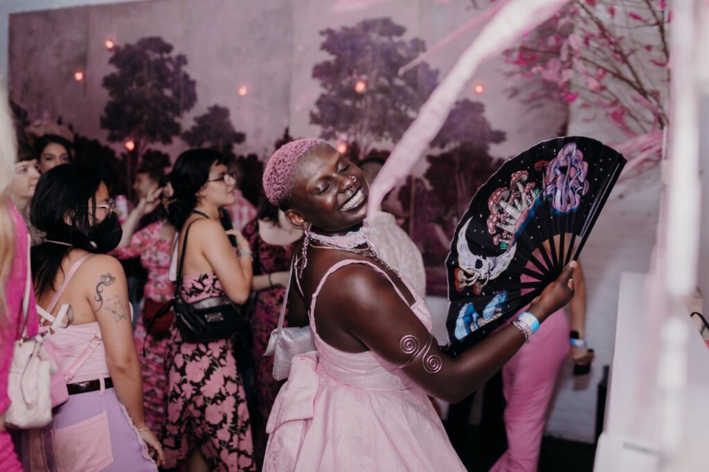 A person with shaved pink hair, a pink dress, and a black fan grins, looking back.