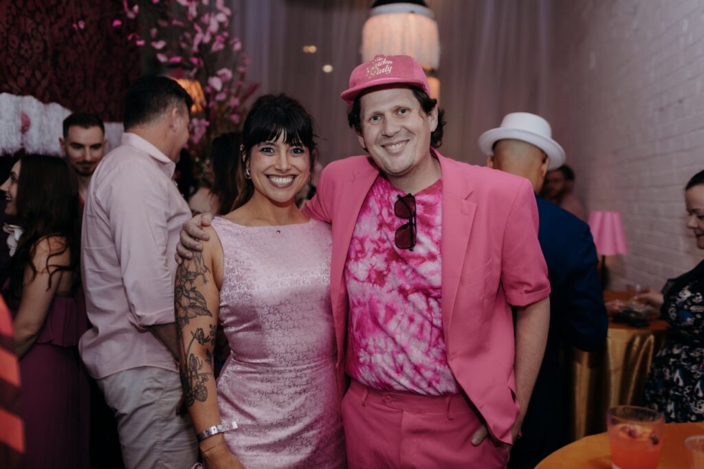 A woman in a pink dress and man in a pink suit jacket, hat, shirt, and pants smile together.
