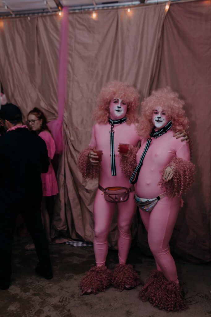 Two people in pink spandex and wigs on their head, hands, and feet stand with drinks.