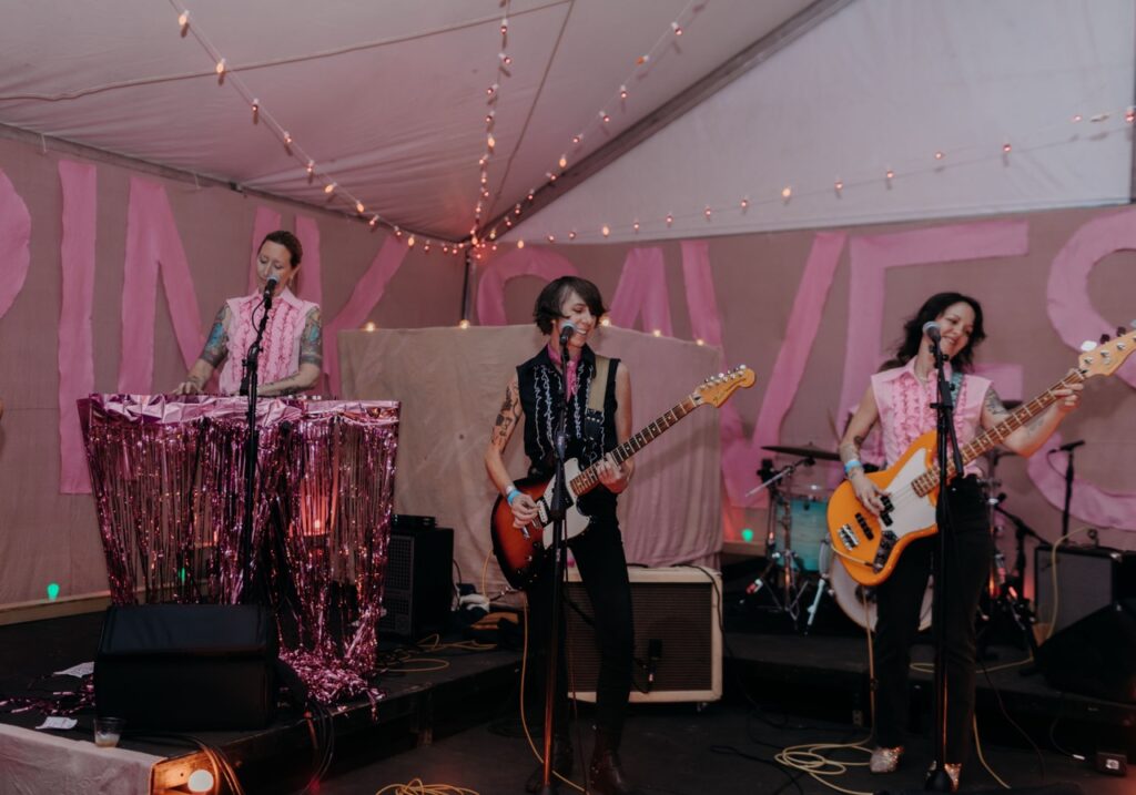 Three women play on stage with pink decor around them.