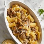 A white serving dish filled with rigatoni pasta and a rich Genovese sauce, topped with grated cheese.