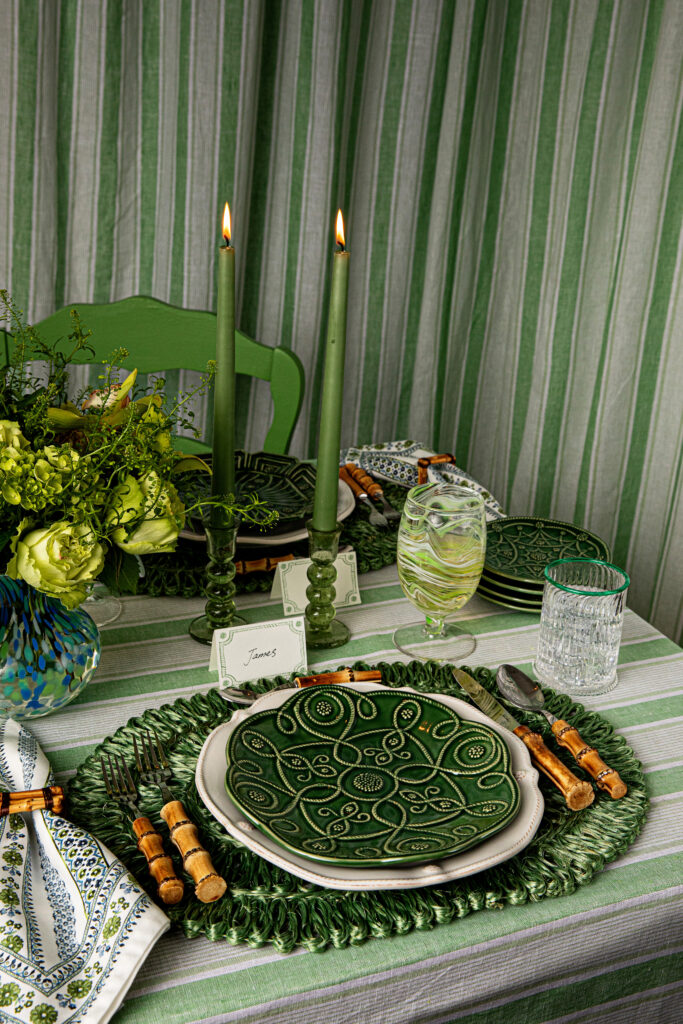 A place setting at a table, with everything in jewel tone green. Candles, the plates, curtain, placemat, etc.
