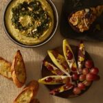 Three appetizers on a linen tablecloth: a bowl of white bean hummus topped with zhoug, endive leaves filled with grapes and cheese, and a piece of bread topped with a dip.