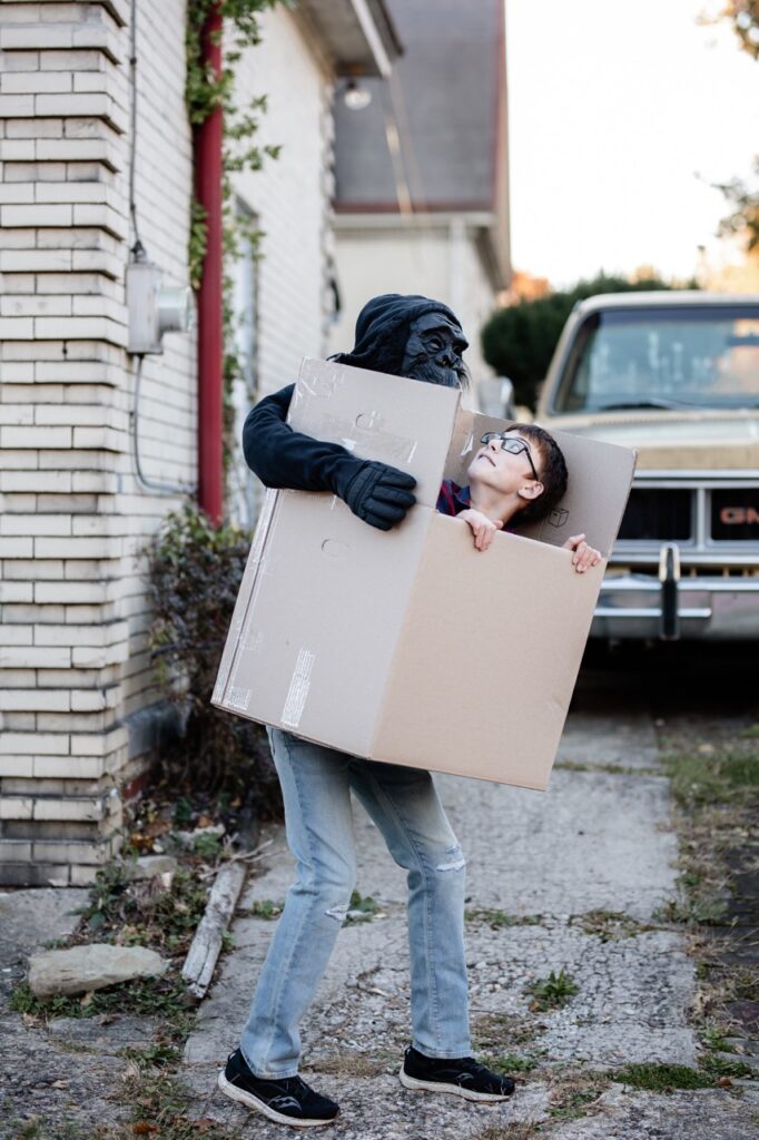 A Halloween costume of a kid in a box being carried by a masked man.