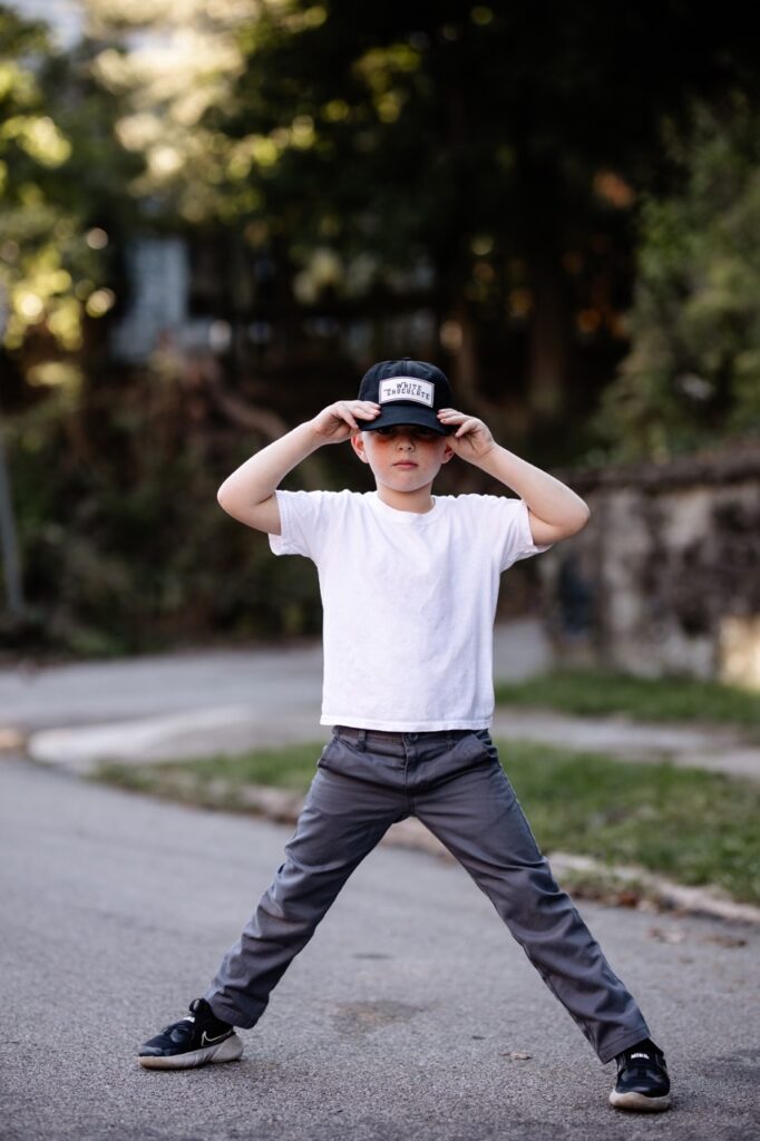 A kid in a white shirt and jeans with a hat stands in the street.