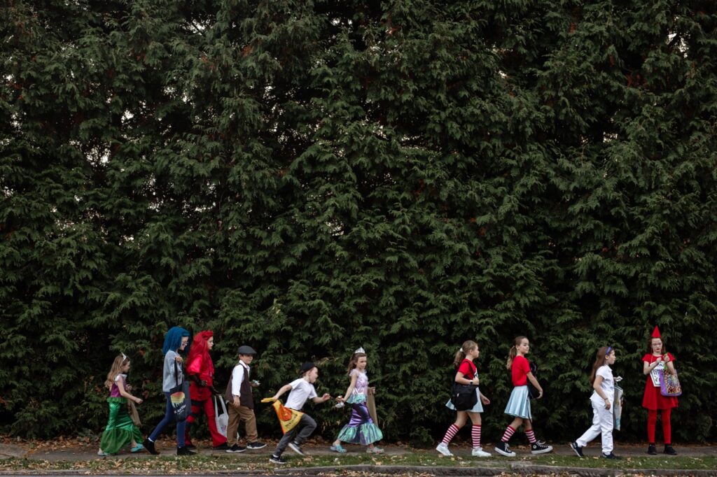 A group of kids in Halloween costumes walk in front of a forest of trees.