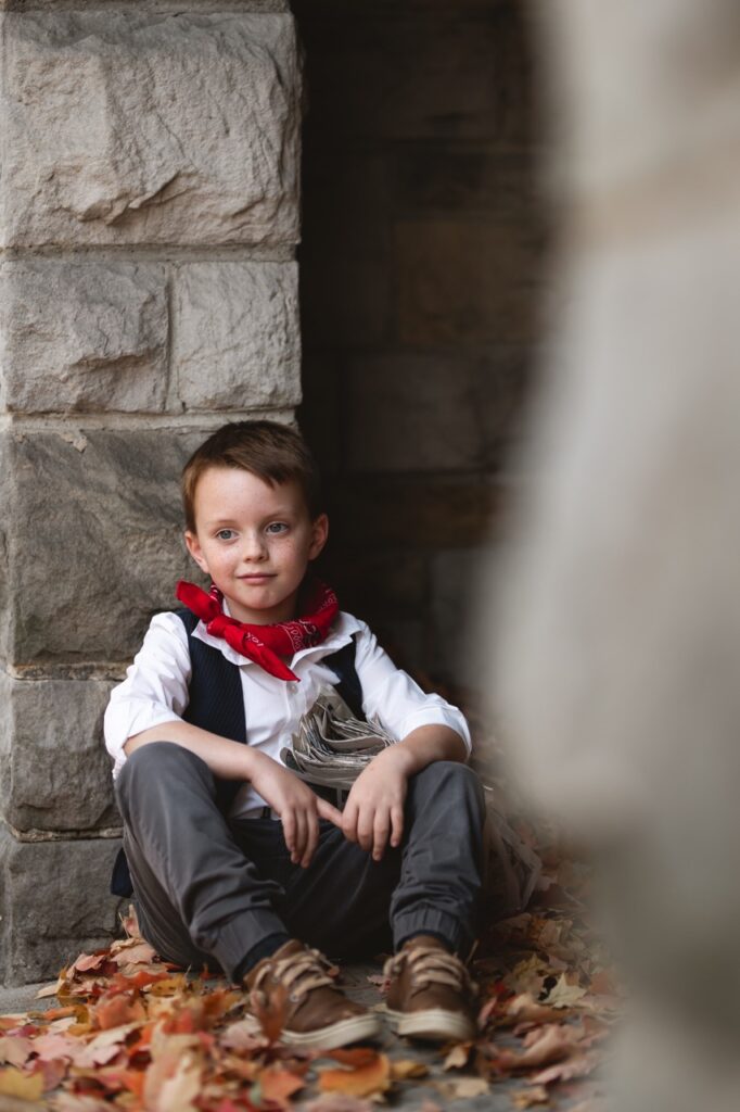 A small boy sits against a stone wall as a cowboy.