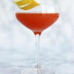 A stemmed glass filled with a pinkish-red Jack Rose cocktail, garnished with a spiral of lemon peel.