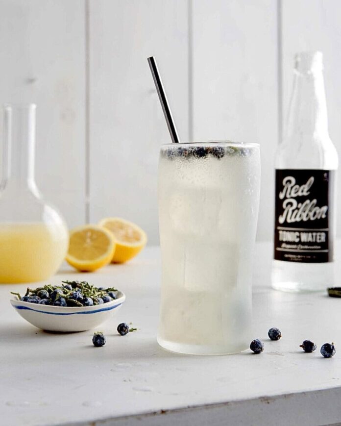 A juniper lemonade tonic sits on a table with juniper berries for a garnish and around it as well as lemons and a bottle of red ribbon soda.
