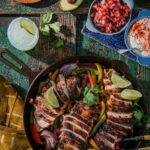 A chicken fajita skillet with slices chicken, limes, and peppers. Then there are small bowls of pico, sour cream, and onions.