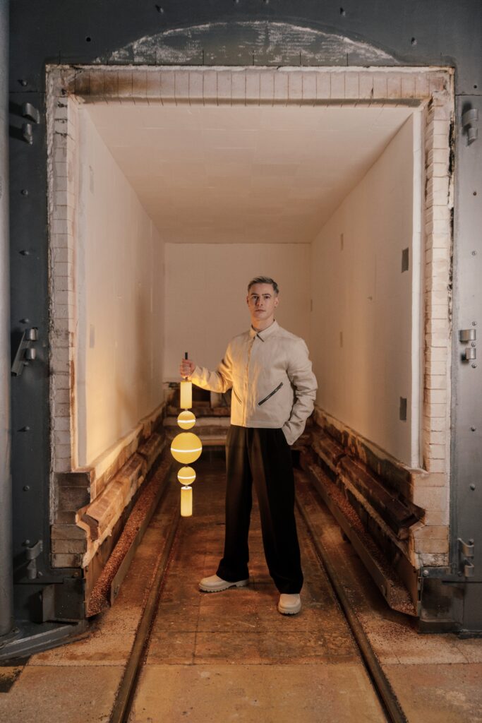 A white male in a cream jacket and black slacks holds a lamp made of balls and cylinders.