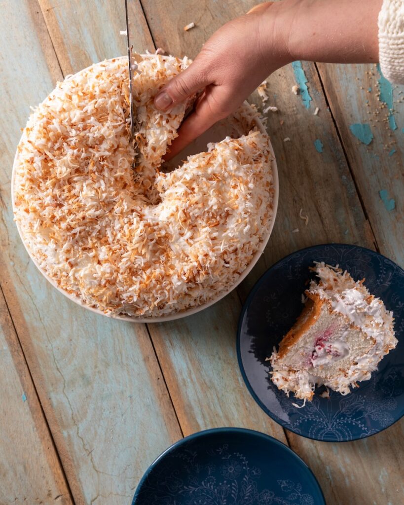 A woman cuts a slice of Martha Stewart-Inspired Angel Food Cake that's topped with coconut as another slice sits on a blue plate nearby.