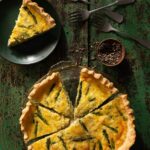 A Martha Stewart-Inspired Asparagus Leek and Jarlsberg Quiche with a slice taken out of it, sitting on a black plate.