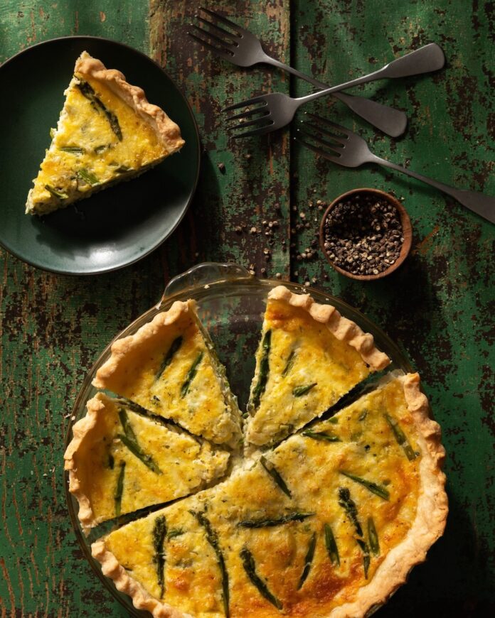 A Martha Stewart-Inspired Asparagus Leek and Jarlsberg Quiche with a slice taken out of it, sitting on a black plate.
