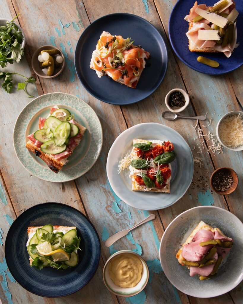 Various light blue and dark blue plates hold different types of focaccia sandwiches like cucumber, cream cheese and salmon, prosciutto, all on a blue and white picnic table.