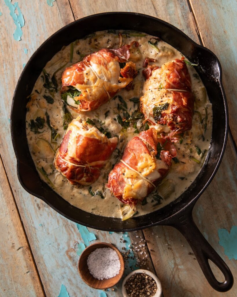 A cast iron pan on a picnic table with chicken roll ups sitting in a creamy sauce with two small containers of salt and pepper below.