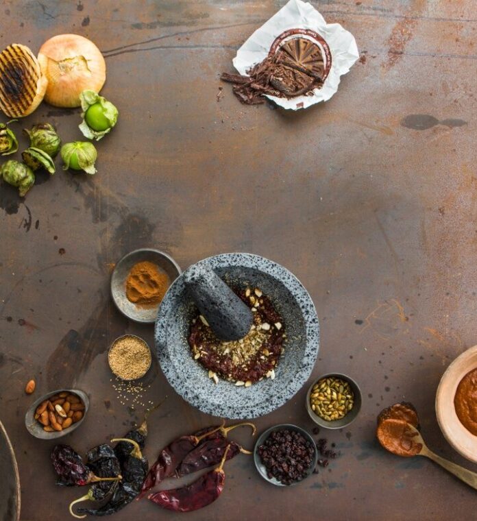 Mexican Mole Sauce A flat lay photo showing ingredients for Mexican mole sauce, including various chiles, nuts, spices, and a stone mortar and pestle on a rustic metal surface.