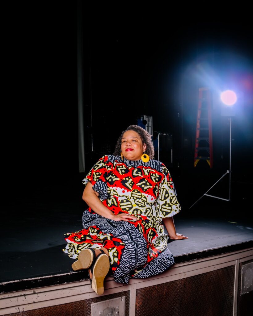 Michele Lee Betts sits on the edge of a stage with a light in the background.
