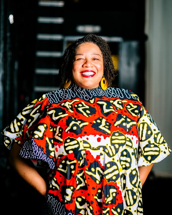 Michele Lee Betts stands in a pattern color dress with a smile on her face.