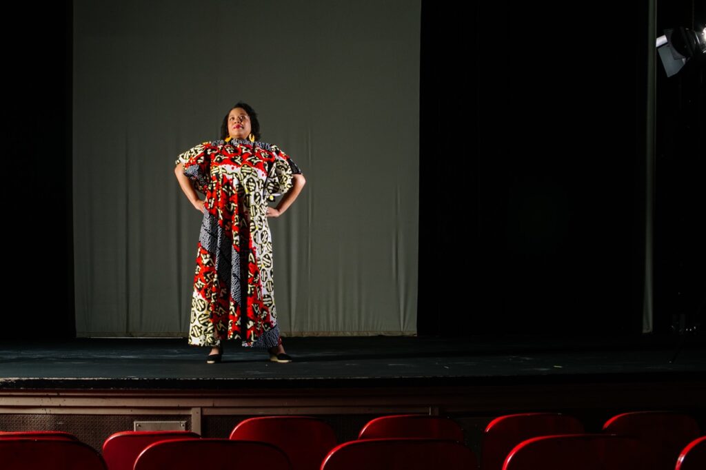 A woman in a long colorful pattern dress stands on a stage with her hands on her hips.