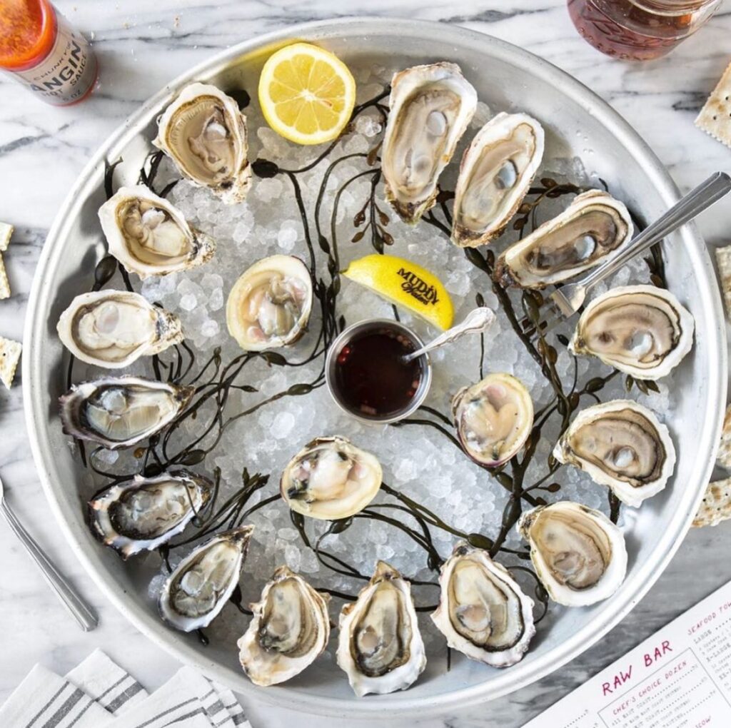 A top-down view of a round platter of raw oysters on the half shell, arranged on a bed of ice and seaweed with a small dish of mignonette, a lemon, and crackers.