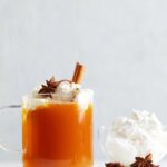 A glass mug of mulled apple cider topped with vegan whipped cream, a cinnamon stick, and a star anise pod.