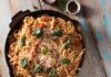 Martha Stewart-Inspired One Pan Pasta In a skillet pan sits a large servings of pasta with tomatoes, basil, and cheese on top. It all sits on a picnic table with forks and a small container of parmesan underneath the pan.