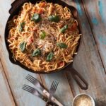 In a skillet pan sits a large servings of pasta with tomatoes, basil, and cheese on top. It all sits on a picnic table with forks and a small container of parmesan underneath the pan.