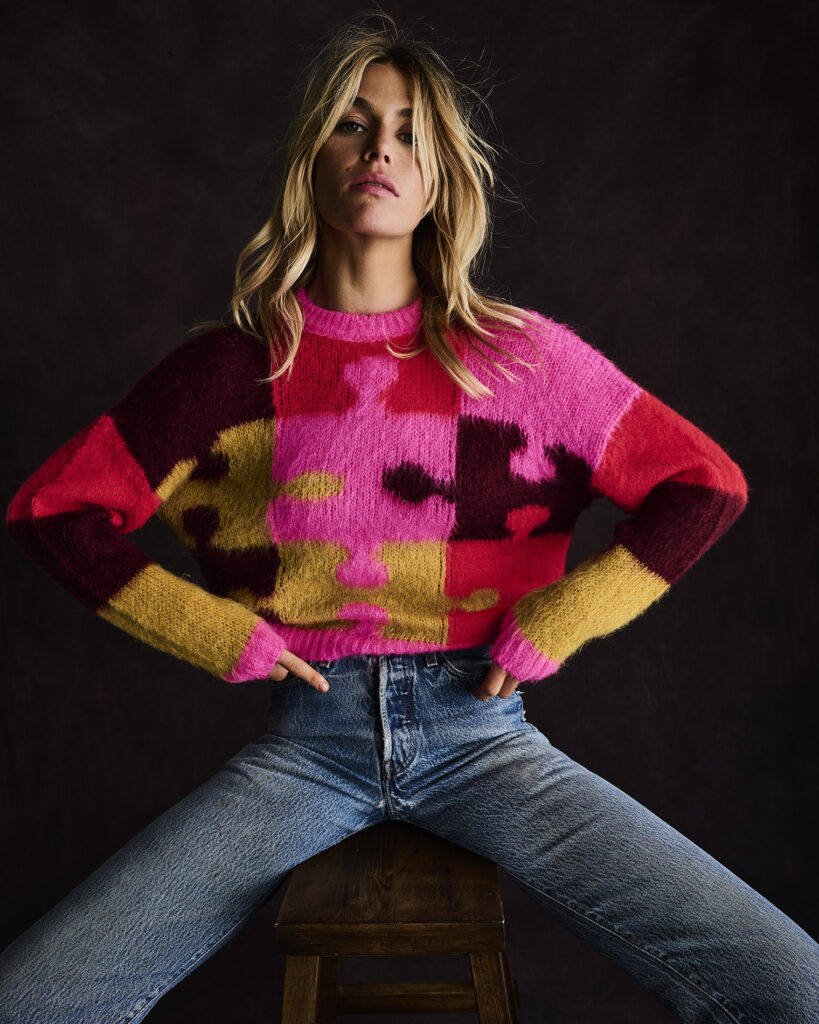 A women in jeans and a brightly colored sweater in a brown, pink, and red puzzle piece pattern sits on a stool.