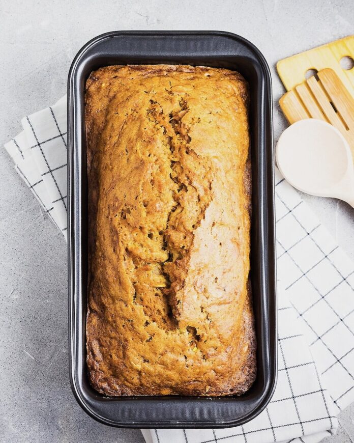 A loaf of Pawpaw Sweet Bread in an orange color sits in a loaf pan on a white towel and counter.