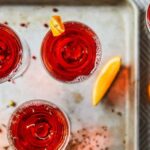 Three Pink Peppercorn Negronis in coupe glasses on a metal tray, garnished with orange peels and scattered pink peppercorns.