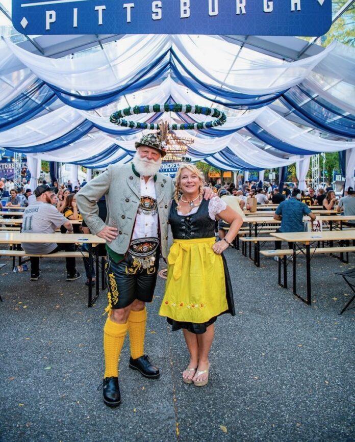 A man and woman stands in front of an Oktoberfest Pittsburgh sign outside.