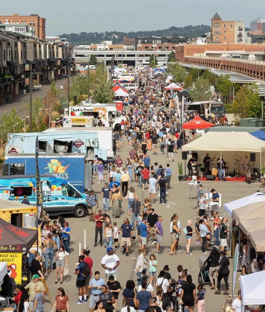 People gather in a crowd in Pittsburgh for the Pittsburgh taco festival, walking down a street of food trucks. 