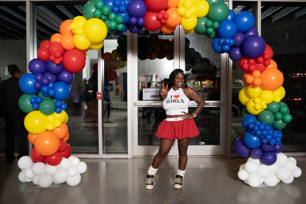 A woman in a white tank top and red skirt holds out a peace sign under a rainbow of balloons.