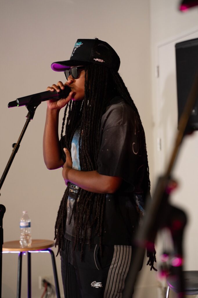 A person with long braids, sunglasses, and a baseball hat sings into a mic.