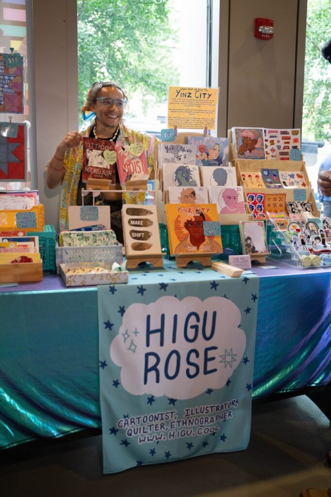 A queer market vendor smiles at their table of books and cards.