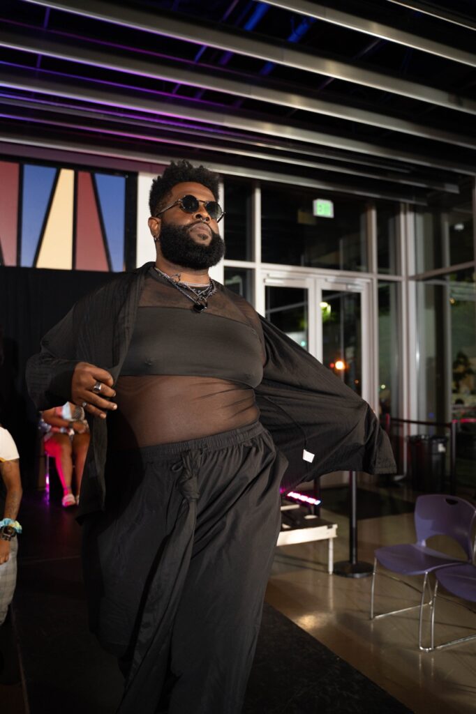 A person in a black top, pants, and sunglasses poses on a catwalk.