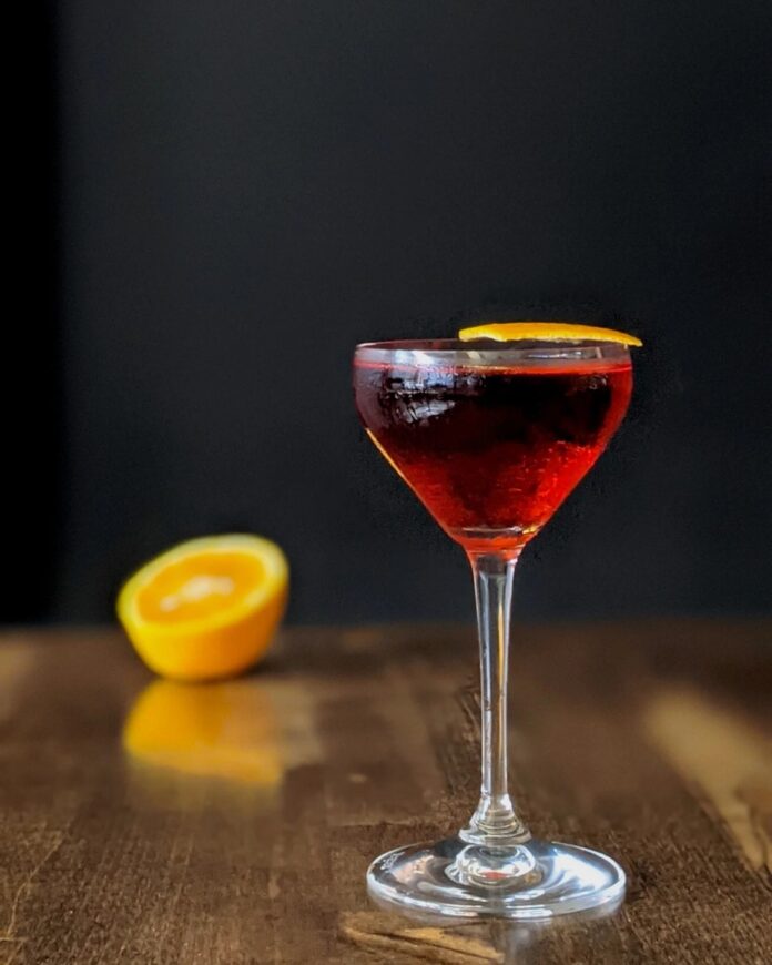Q-groni A cocktail in a stemmed glass with an orange peel garnish, with a blurred orange half in the background.