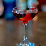A red cocktail in a coupe glass with an orange peel garnish, with a blurry background of bar bottles.