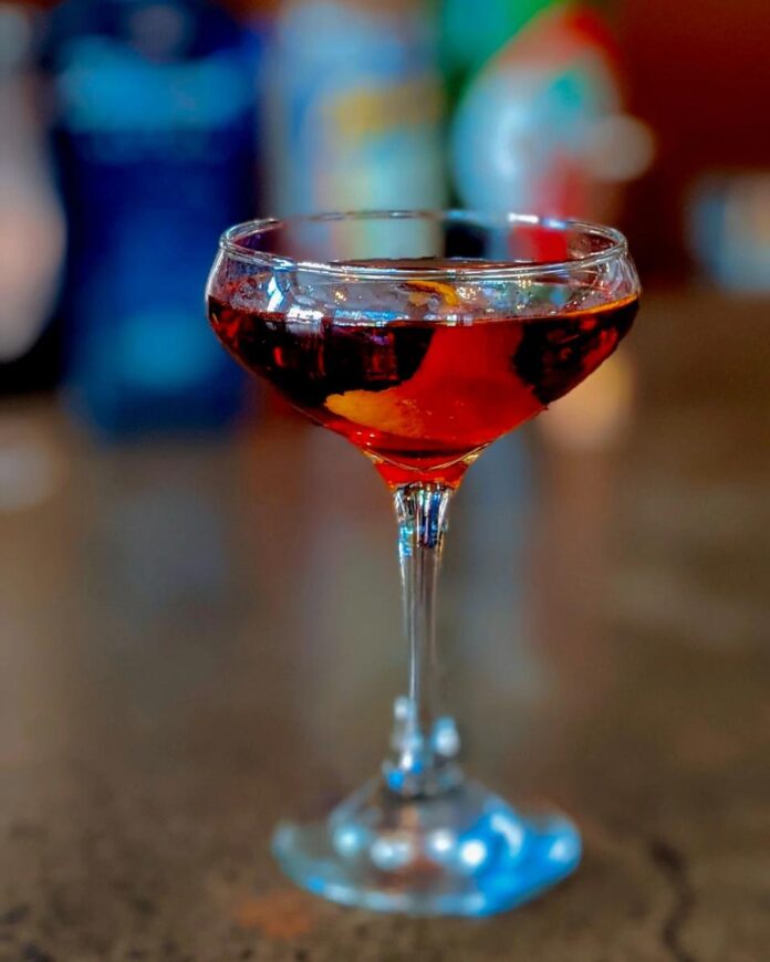 Red Wedding A red cocktail in a coupe glass with an orange peel garnish, with a blurry background of bar bottles.
