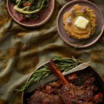 A table set with dishes of food, including a platter of red wine-braised lamb shanks, a plate of braised greens, and a bowl of parsnip mash.