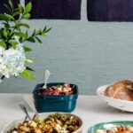 Three bowls of food on it: a roasted beets salad, a brussels sprouts dish, and a carrot dish, with a bouquet of flowers and a loaf of bread nearby.