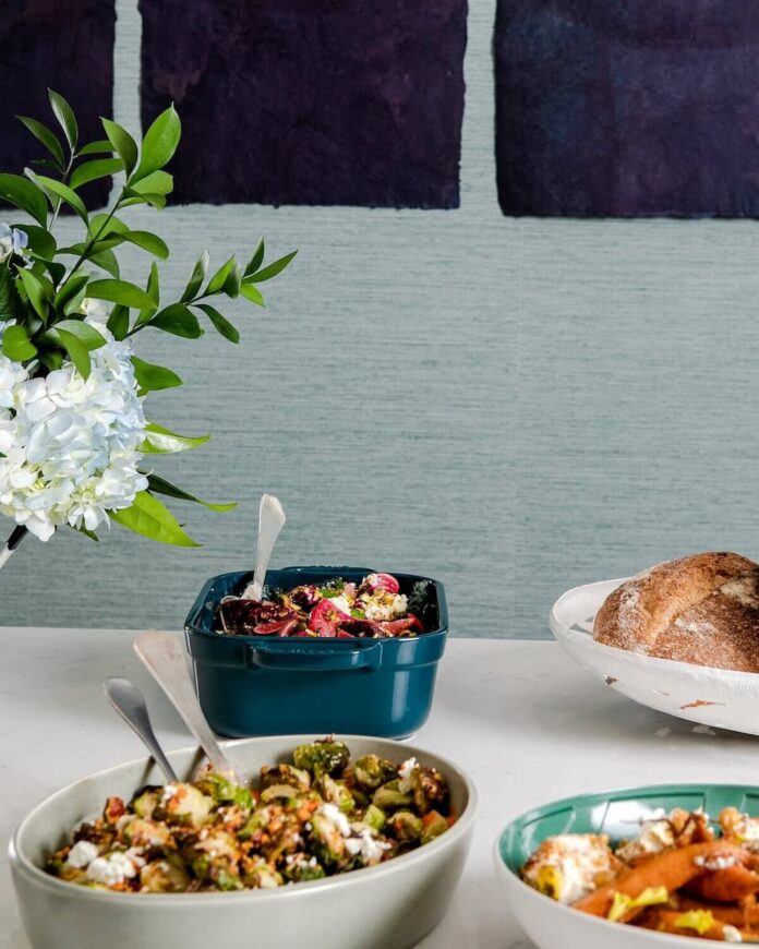 Three bowls of food on it: a roasted beets salad, a brussels sprouts dish, and a carrot dish, with a bouquet of flowers and a loaf of bread nearby.