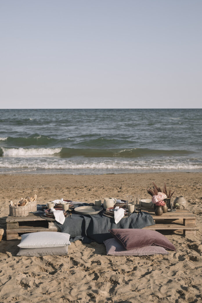 A short picnic table along with pillows and blankets sit on the sand with a wave coming in in the distance.