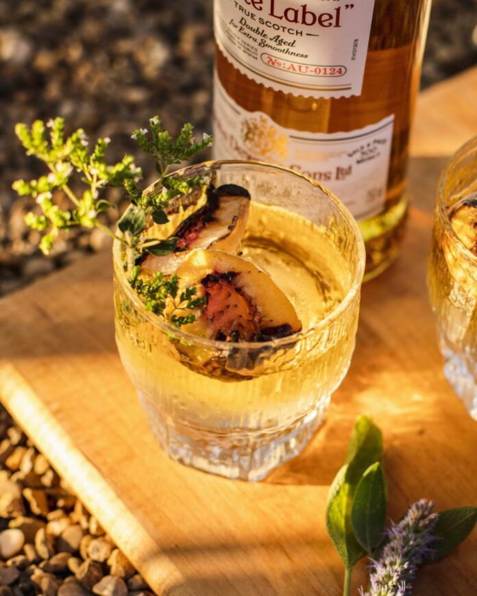 A Smoked Maple and Scotch Old Fashioned cocktail in a rocks glass, garnished with grilled peaches and fresh herbs, with a bottle of Scotch in the background.